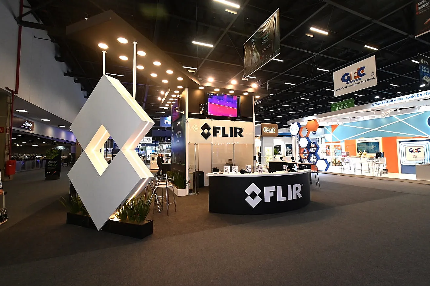 Stand da Flir, com totem em losango iluminado, balcão curvo preto e painel de câmeras termográficas em exposição.