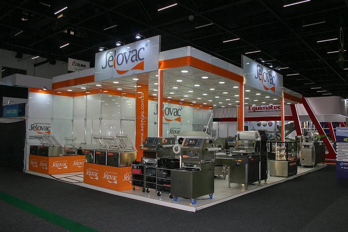 Stand padrão modular com testeiras laranja e branca, equipamentos expostos e iluminação embutida.
