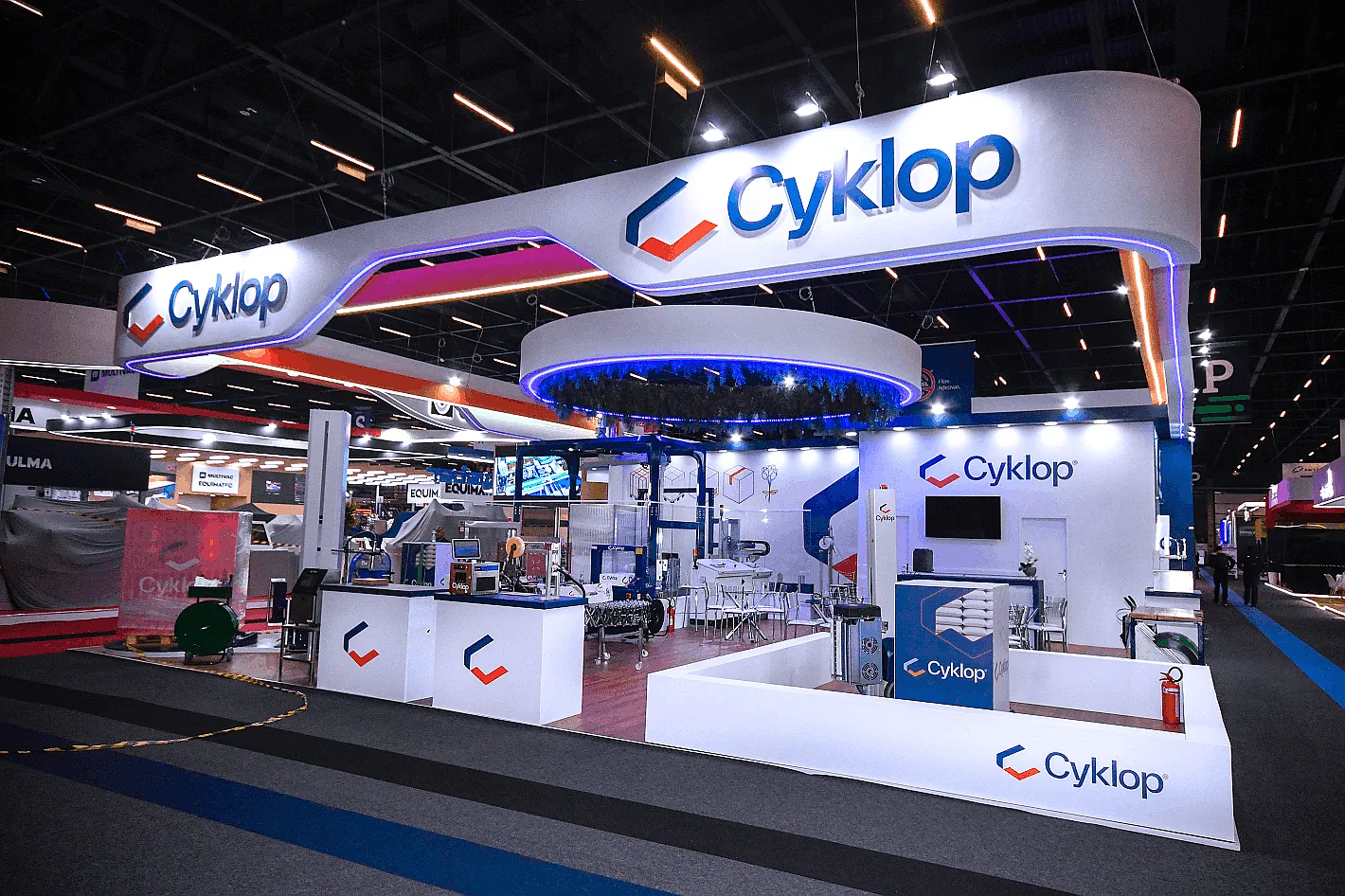 Stand da Cyklop em ilha, com grande testeira curva suspensa, anel de LED azul e equipamentos industriais em operação.