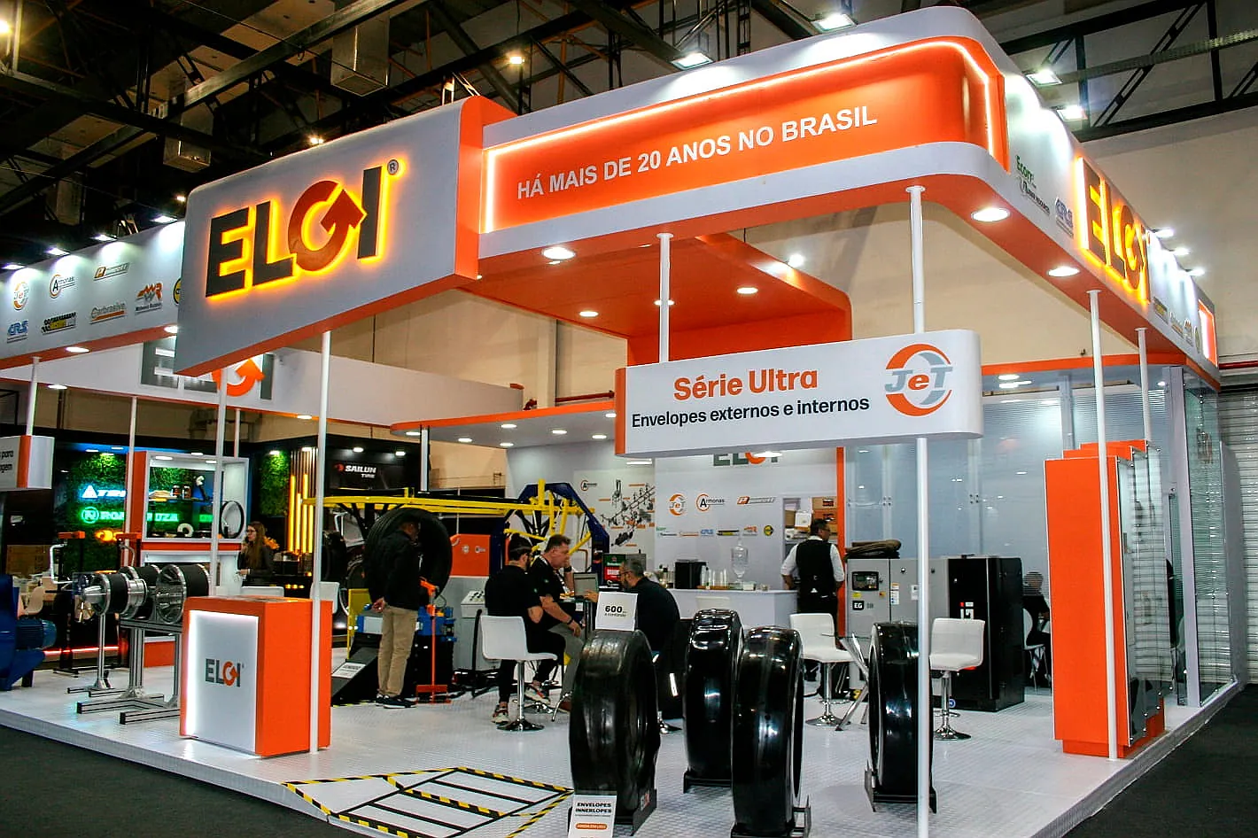 Stand da Eloi, com testeira suspensa iluminada em laranja, lounge de atendimento e produtos automotivos em exposição.