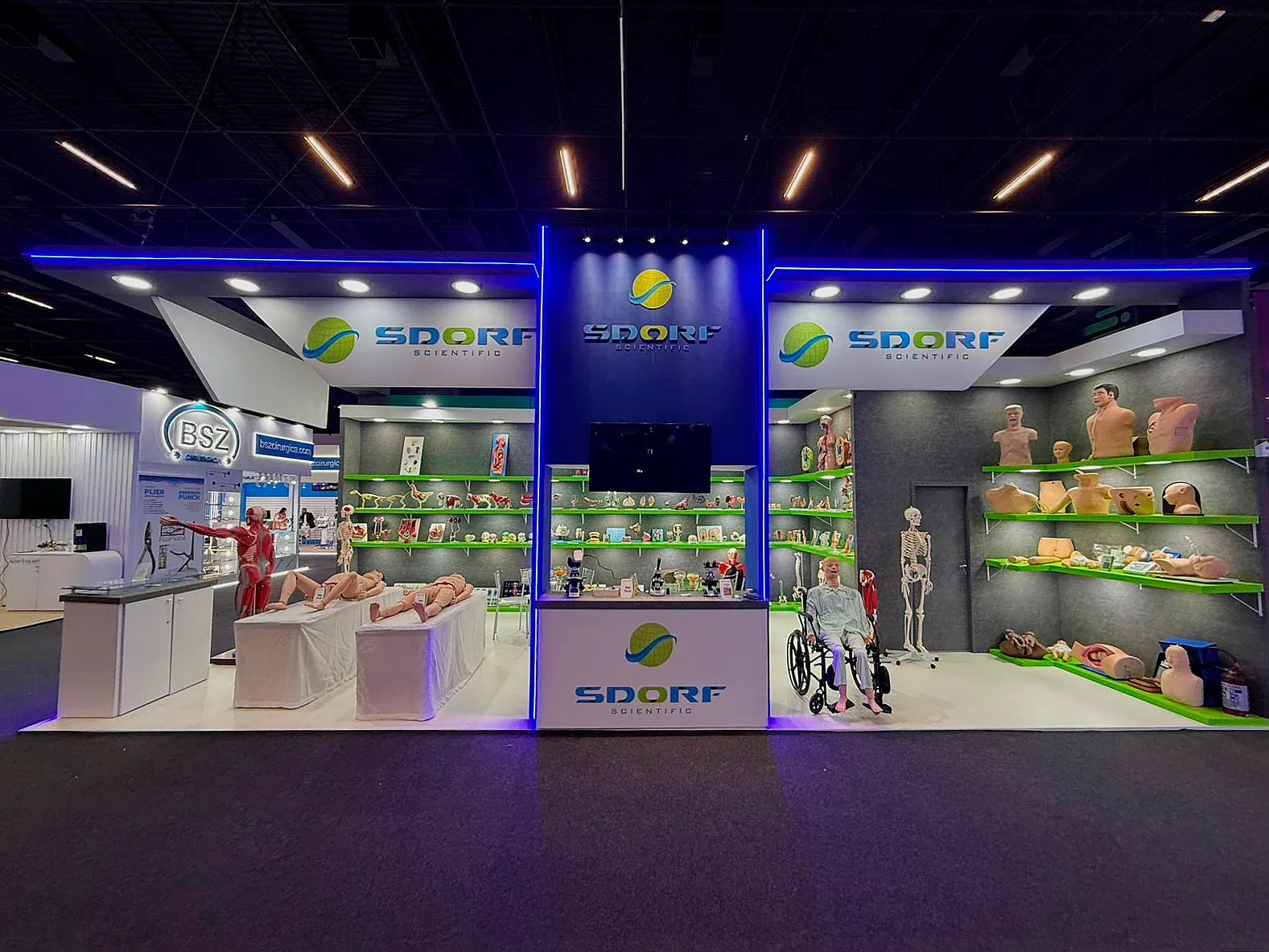 Stand da Sdorf Scientific na Feira Hospitalar, com fita de LED azul, prateleiras verdes e modelos anatômicos em exposição.