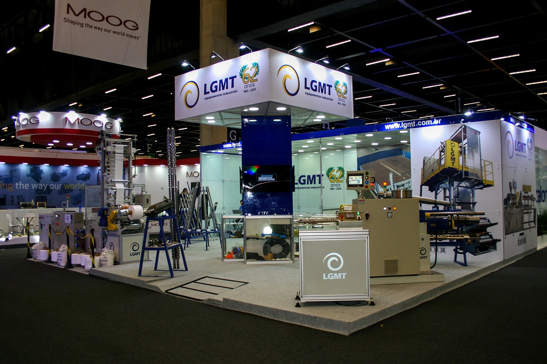 Stand da LGMT na Plástico Brasil, com testeira suspensa de 62 anos, equipamentos industriais em operação e fechamento em vidro.