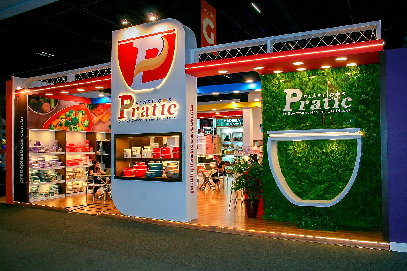 Stand da Pratic Plásticos, com totem de marca iluminado, prateleiras de utilidades domésticas e parede verde natural.