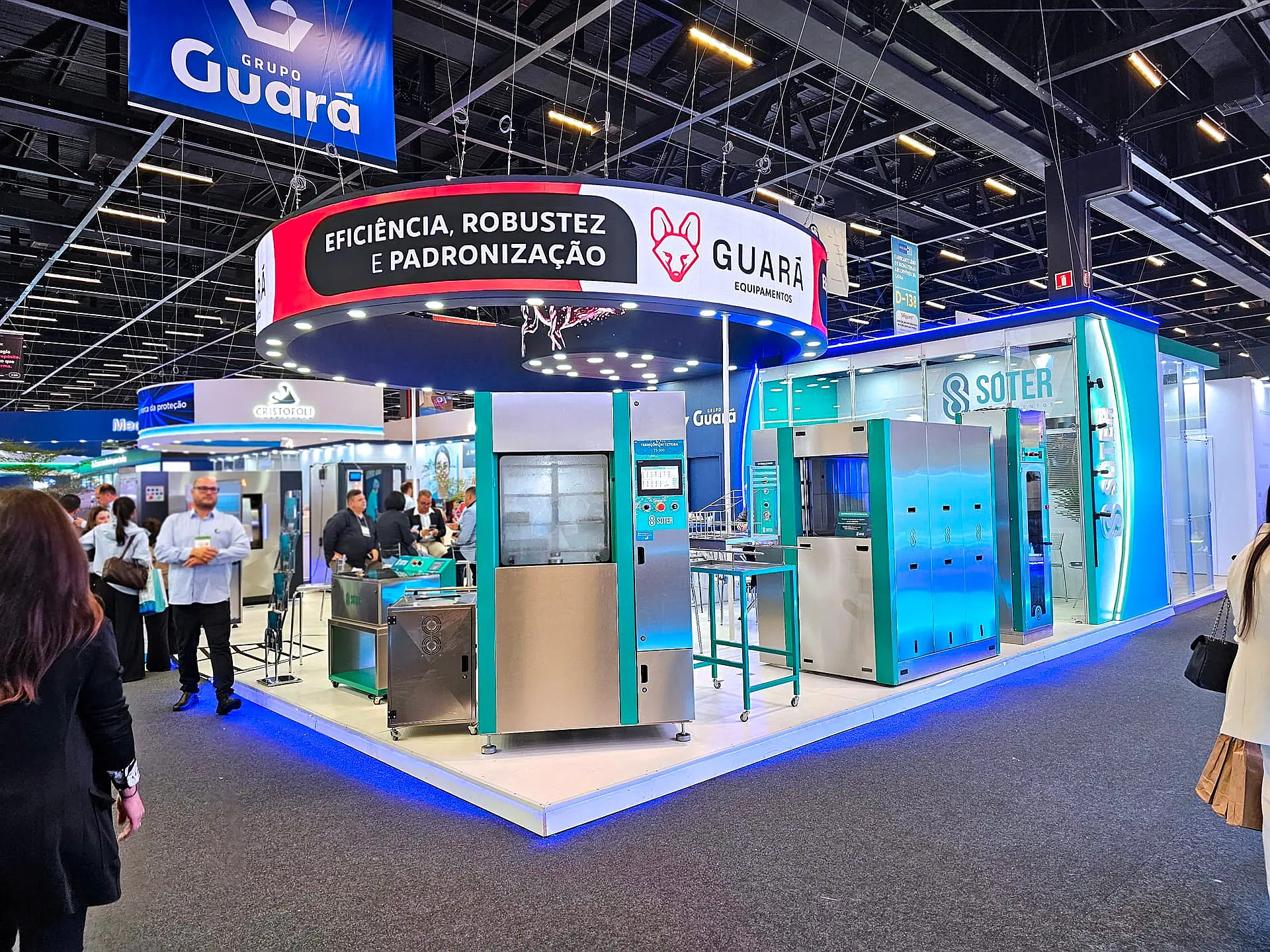 Segundo ângulo de stand em ilha com testeira curva, iluminação perimetral e equipamentos industriais na Feira Hospitalar 2026.