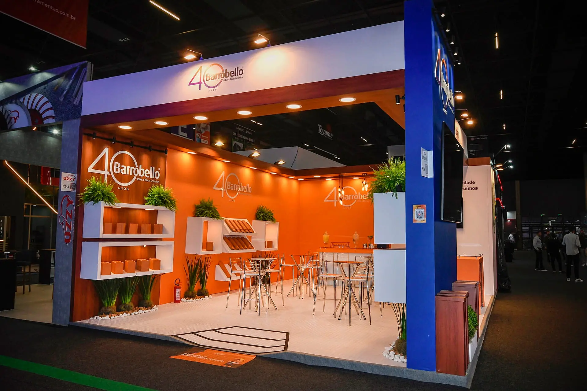 Stand construído com painéis alaranjados, prateleiras de exposição de produtos e área de café na Feicon 2026.