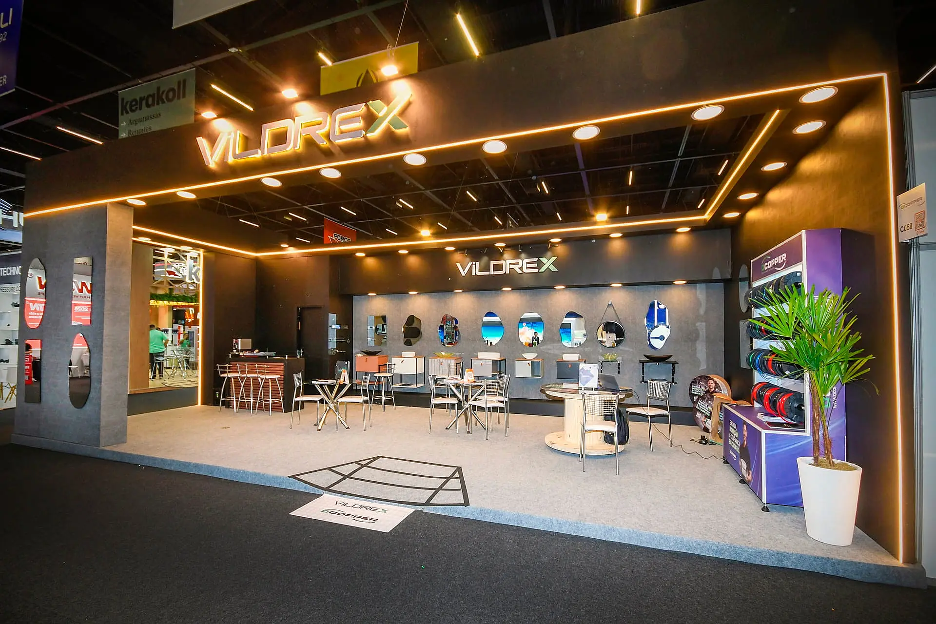 Stand construído em textura cinza, com sanca iluminada em LED âmbar e lounge de mesas altas na Feicon 2026.