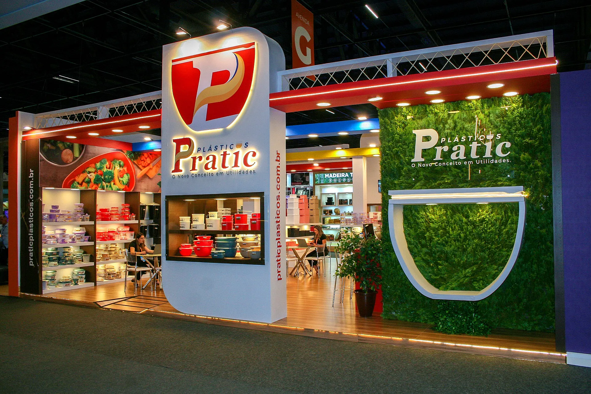 Stand da Pratic Plásticos na Abup, com torre de logotipo iluminada, parede verde natural e prateleiras de potes e utilidades domésticas.