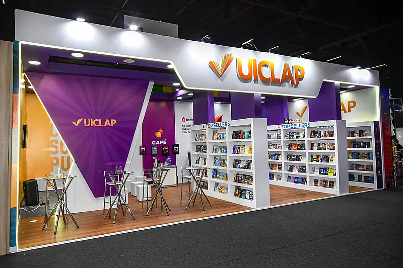 Stand construído roxo e branco com piso de madeira, estantes de livros, lounge e área de café.